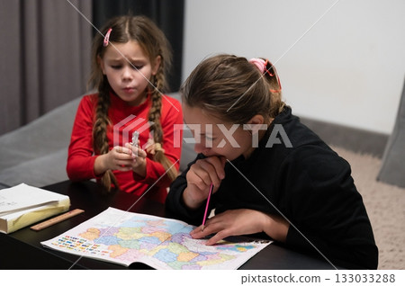 Two young girls are engrossed in studying a colorful map together, sharing a quiet moment of learning and discovery, indoors on a table. Two young girls are engrossed in studying a colorful map together, sharing a quiet moment of learning and discovery, indoors on a table. 133033288