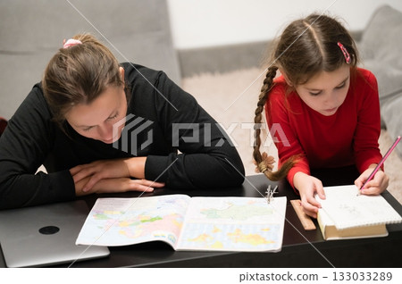Mother and daughter studying together at home. Focusing on homework and education with map and notebook on table, creating family learning time. Mother and daughter studying together at home. Focusing on homework and education with map and notebook on table, creating family learning time. 133033289