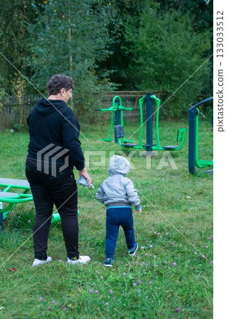 Woman and child walk in a park with exercise equipment. Trees line the background of a grassy area with purple flowers. Woman and child walk in a park with exercise equipment. Trees line the background of a grassy area with purple flowers. 133033512