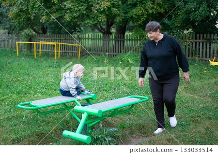 A baby sits on a green exercise bench with an adult walking nearby, showcasing intergenerational activity in an outdoor space. 133033514