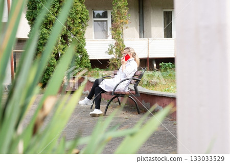 Female doctor in lab coat sits on a park bench talking on the phone in front of a clinic. She is wearing casual clothes. Female doctor in lab coat sits on a park bench talking on the phone in front of a clinic. She is wearing casual clothes. 133033529