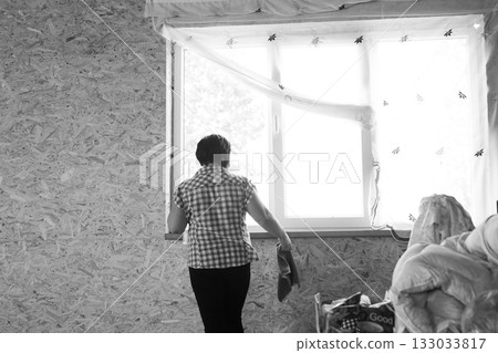 Woman cleaning a window in room with walls of pressboard, sunlight streams in. A glimpse of domestic life, captured in monochrome. Woman cleaning a window in room with walls of pressboard, sunlight streams in. A glimpse of domestic life, captured in monochrome. 133033817