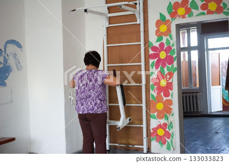 A woman adds an attachment to white and wood stall bars in a room with flower paintings adorning the wall near a doorway. 133033823