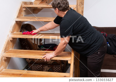 A woman wiping down the wooden stairs in her house, cleaning with a cloth in hand, keeping her home neat and tidy, while wearing gloves. 133033827