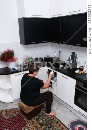 Woman cleaning under the sink in her kitchen, using cleaner in the white and black kitchen. An interesting array of rugs on the floor. 133033831