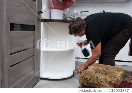 Woman cleaning a kitchen cabinet with a cleaning spray. The cabinet is white and has two shelves. There is a door and a stool nearby. 133033837