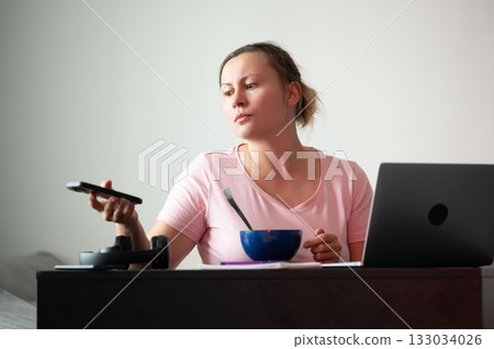 Woman working from home, holding phone while eating. A laptop is next to a bowl of food on a dark desk during a casual day indoors. 133034026