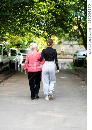 Walking through the park, a young woman supports an elderly lady as they enjoy a warm afternoon together amidst the trees and parked cars 133034170