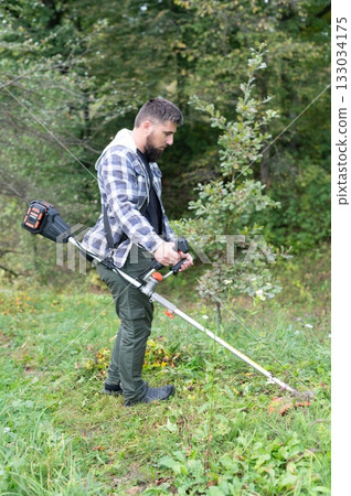 Man trimming grass in a yard with a string trimmer. Yard work and landscaping with battery powered yard tools for lawn maintenance. 133034175