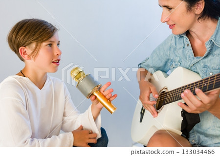A young musician practices singing into a microphone while their instructor plays acoustic guitar. A collaborative music lesson. A young musician practices singing into a microphone while their instructor plays acoustic guitar. A collaborative music lesson. 133034466