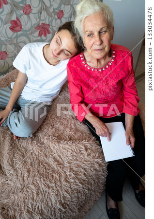 Elderly woman and young girl share a tender moment of connection while sitting together on a cozy bed surrounded by soft textures in a warmly lit room Elderly woman and young girl share a tender moment of connection while sitting together on a cozy bed surrounded by soft textures in a warmly lit room 133034478