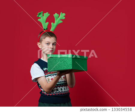 Happy boy with green antlers holds stack of red and green Christmas gift boxes, smiling in front of red background in festive clothes Happy boy with green antlers holds stack of red and green Christmas gift boxes, smiling in front of red background in festive clothes 133034558