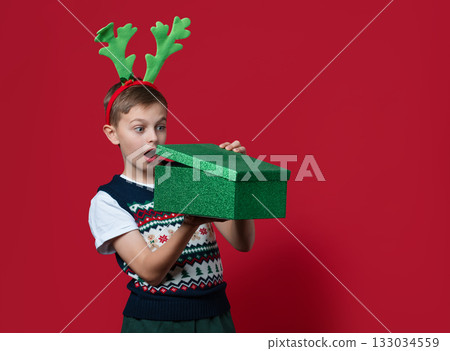 Happy boy with green antlers holds stack of red and green Christmas gift boxes, smiling in front of red background in festive clothes 133034559