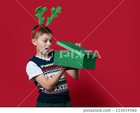 A surprised boy in holiday sweater and green reindeer antlers opening 133034560