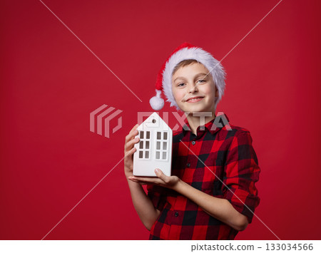 Merry Christmas. A smiling boy in red plaid shirt and Santa hat holding a white ceramic house, festive mood on red background 133034566