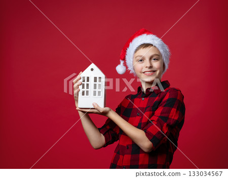 Boy in red plaid shirt Santa hat holding white ceramic house, festive mood on red background. Santa holds gift to house. New Year's gift. Boy in red plaid shirt Santa hat holding white ceramic house, festive mood on red background. Santa holds gift to house. New Year's gift. 133034567