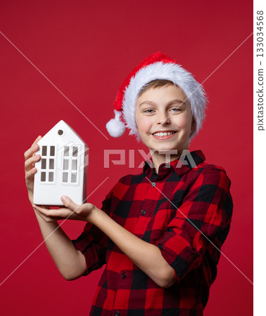 Boy in red plaid shirt Santa hat holding white ceramic house, festive mood on red background. Santa holds gift to house. New Year's gift. Boy in red plaid shirt Santa hat holding white ceramic house, festive mood on red background. Santa holds gift to house. New Year's gift. 133034568