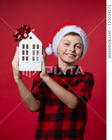 Merry Christmas. A smiling boy in red plaid shirt and Santa hat holding a white ceramic house, festive mood on red background Merry Christmas. A smiling boy in red plaid shirt and Santa hat holding a white ceramic house, festive mood on red background 133034571