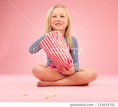 Popcorn, snack and happy girl in a studio with pink background sitting with movie snacks. Food, happiness and hungry young child with a paper bag and chips eating and feeling relax with a smile 133034702