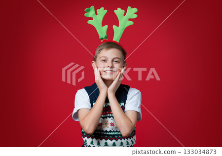 Excited boy festive vest and green reindeer antlers holding his face in surprise, joyful expression, red background Excited boy festive vest and green reindeer antlers holding his face in surprise, joyful expression, red background 133034873