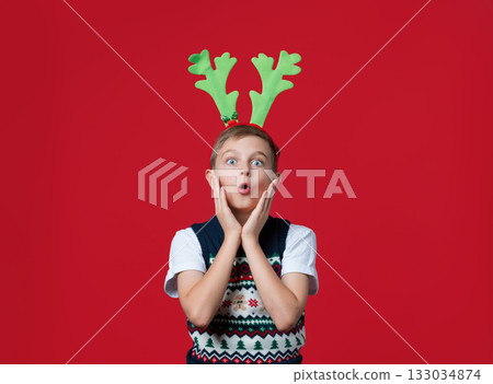 Excited boy in festive vest and green reindeer antlers holding his face in surprise Excited boy in festive vest and green reindeer antlers holding his face in surprise 133034874