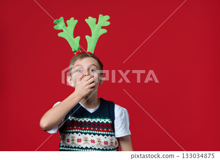 Excited boy festive vest and green reindeer antlers holding his face in surprise, joyful expression, red background Excited boy festive vest and green reindeer antlers holding his face in surprise, joyful expression, red background 133034875