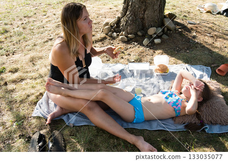 A mother and daughter enjoy a relaxing picnic by a tree, sharing a meal and enjoying the summer day. 133035077