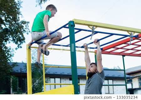 Father and son enjoy an active day at the playground, climbing and exercising together. Father and son enjoy an active day at the playground, climbing and exercising together. 133035234