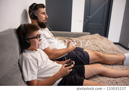 A father and son bond over a gaming session, comfortably lounging on a couch with headphones and controllers. A father and son bond over a gaming session, comfortably lounging on a couch with headphones and controllers. 133035237
