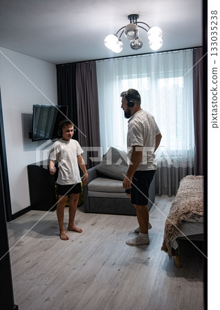Father and son, both wearing headphones, stand facing each other in a living room, seemingly ready to dance or engage in a playful activity. 133035238