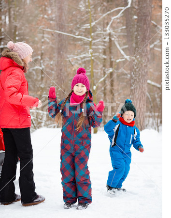 Children and their mom play together in fresh snow, dressed in warm colorful winter clothes, surrounded by trees in winter wonderland. 133035270