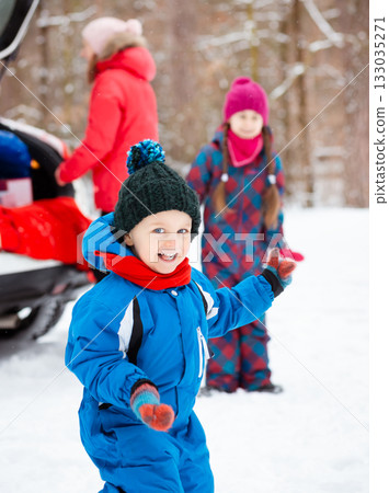 A cheerful young boy dressed in blue snowsuit and black hat plays in the snow, smiling with trees and family in the background. 133035271