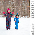 Children and their mom play together in fresh snow, dressed in warm colorful winter clothes, surrounded by trees in winter wonderland. 133035304