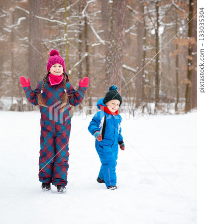 Children and their mom play together in fresh snow, dressed in warm colorful winter clothes, surrounded by trees in winter wonderland. 133035304