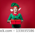Happy boy dressed as surprise , impression, emotions Christmas elf holding a red present with ribbon, green red costume with pointy hat and elf ears 133035586