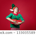 Happy boy dressed as surprise , impression, emotions Christmas elf holding a red present with ribbon, green red costume with pointy hat and elf ears 133035589