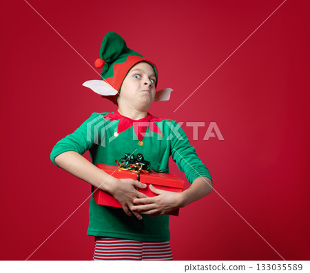 Happy boy dressed as surprise , impression, emotions Christmas elf holding a red present with ribbon, green red costume with pointy hat and elf ears Happy boy dressed as surprise , impression, emotions Christmas elf holding a red present with ribbon, green red costume with pointy hat and elf ears 133035589