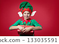 Merry Christmas concept. Smiling funny boy in Santa's elf costume holding Christmas gift in his hand. Shooting on red background. 133035591