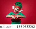 Merry Christmas concept. Smiling funny boy in Santa's elf costume holding Christmas gift in his hand. Shooting on red background. 133035592