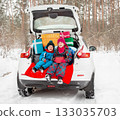 Merry Christmas concept. Two children in colorful winter suits open Christmas presents while sitting in trunk of white car surrounded 133035703