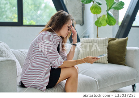 Sad serious middle aged woman with smartphone sitting on sofa Sad serious middle aged woman with smartphone sitting on sofa 133037255