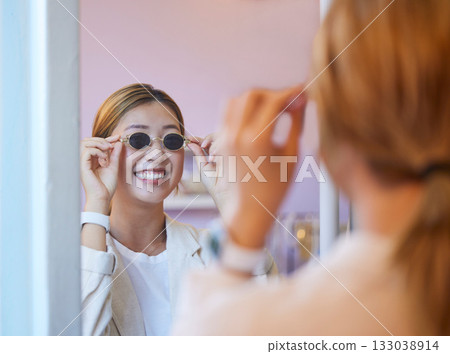 Retail, fashion and sunglasses with Asian woman in mirror for boutique, consumer and designer eyewear. Happy, smile and shopping with girl customer in store for beauty, luxury and discount deal 133038914