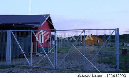 Red farmhouse behind white metal gates and orange building in the distance, farm provincial landscape with residential and outbuildings, no people, farmer's housing in the countryside 133039000