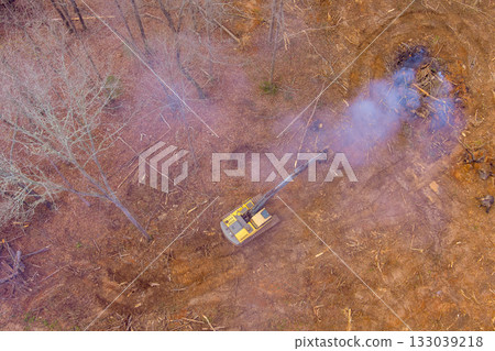 Heavy machinery clears forested area while smoke rises from burning debris 133039218