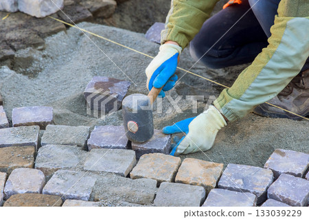 Worker laying cobblestones for pathway construction in residential area during work day hours Worker laying cobblestones for pathway construction in residential area during work day hours 133039229