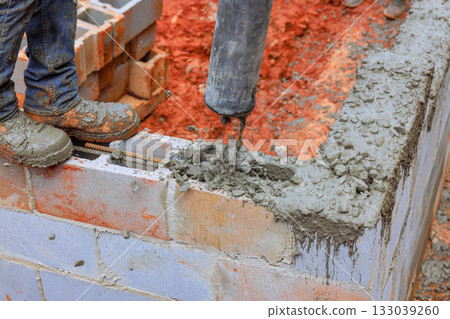 Construction worker pouring concrete into cinder block wall at building site during daylight hours 133039260