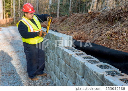 Construction worker measures stone blocks wall building along gravel pathway during work day hours Construction worker measures stone blocks wall building along gravel pathway during work day hours 133039386