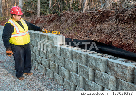 Construction worker checks level of stone retaining wall along wooded area during daylight hours Construction worker checks level of stone retaining wall along wooded area during daylight hours 133039388