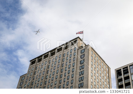 Tall office building with national flag on rooftop and airplane flying in cloudy blue sky Tall office building with national flag on rooftop and airplane flying in cloudy blue sky 133041721