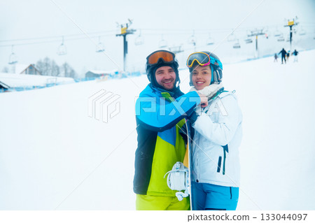 Couple enjoys a fun skiing day on a snowy mountain slope during winter with ski lifts in the background Couple enjoys a fun skiing day on a snowy mountain slope during winter with ski lifts in the background 133044097